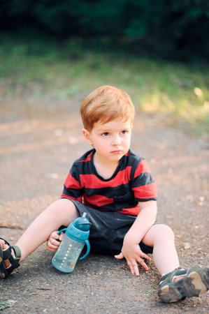 Young child in casual clothes sits on the ground in the summer in the park and drinks water from a drinking cupの写真素材