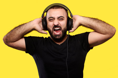 Screaming arabic man with beard listens to music with headphones on yellow isolated background....