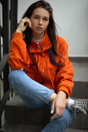 Young trendy hipster woman sits on a summer staircase in a red jacket and blue jeansの写真素材