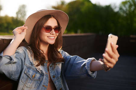 Smiling caucasian attractive woman in straw hat takes a selfie on her smartphone outdoors in summer...
