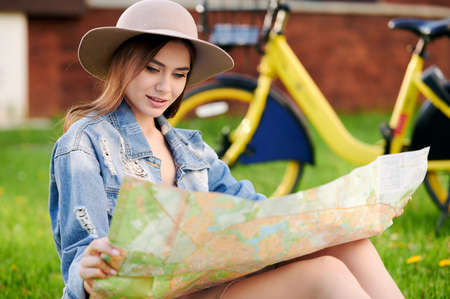 Young hipster tourist woman wearing hat and jeans jacket sits on the grass after cycling and using...