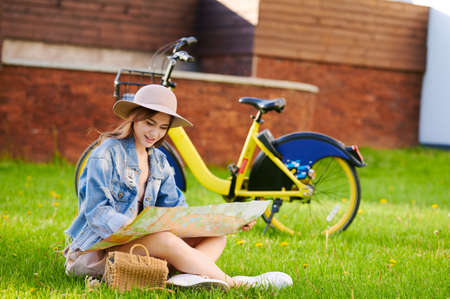 Young woman sitting on the grass in public park and uses paper city map in front of rental bike