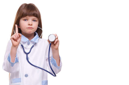 Cute serious little girl doctor wearing white coat holds stethoscope and shows finger gesture on white isolated backgroundの写真素材