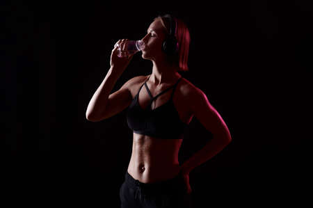 Slim attractive woman in sportswear with headphones drink water after workout on black isolated backgroundの写真素材