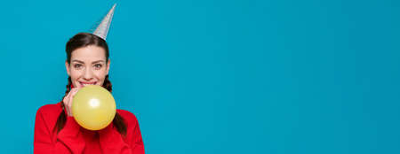 Smiling woman in festive paper cone on her head inflates yellow balloon on blue isolated backgroundの写真素材