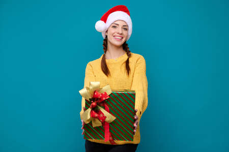 Beautiful young smiling woman in santa claus hat holding a new year gift on a blue isolated...