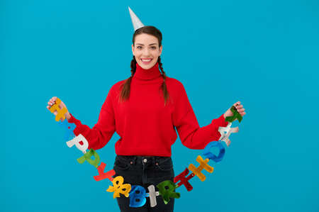 Smiling young woman in celebration cone holding paper letters happy birthday on blue isolated backgroundの写真素材