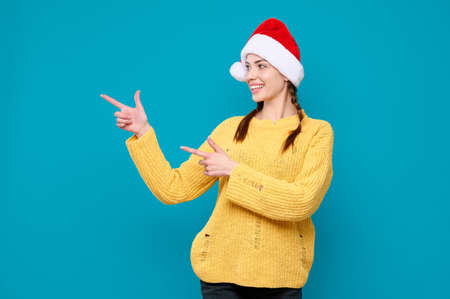 New Year advertising. Young woman in santa claus hat smiling and pointing finger at product on blue isolated backgroundの写真素材