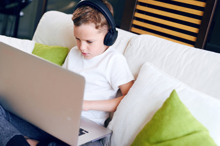 Concentrated schoolboy wearing headphones with laptop play online games at home. Distance learning...