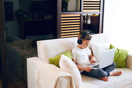Concentrated schoolboy wearing headphones with laptop play online games at home. Distance learning in quarantineの写真素材