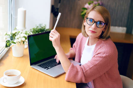 Young beautiful caucasian woman in glasses works at a laptop holds a pen in her hands drinks coffee home office work during quarantineの写真素材
