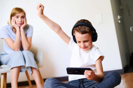 Child boy with mother holds phone is lying on the floor with headphones watching cartoons, listening...