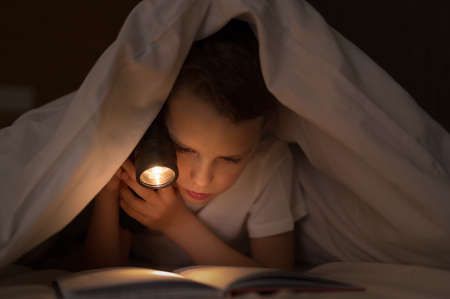 Caucasian child boy reading interesting book under the covers at nightの写真素材
