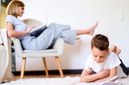 Son reads book on the floor while mom works at laptop sitting on an armchair. Distance learning...