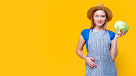Woman gardener holding white cabbage in hands and smiling on yellow isolated studio backgroundの写真素材