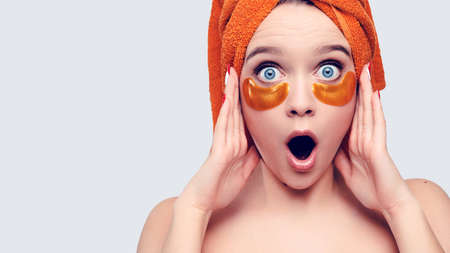 Surprised young caucasian woman with patches under her eyes and a towel on her head holds card product . High quality photoの写真素材