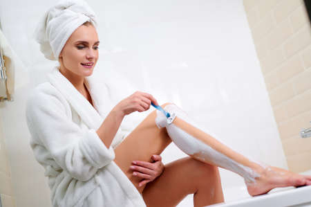 Caucasian woman shaving legs with razor blade in bathroom while sitting at the edge of the bathtub....