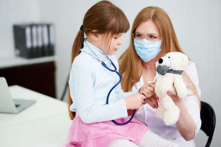 Caucasian female doctor wear face mask show little girl examining bear toy by stethoscope indoor hospitalの写真素材