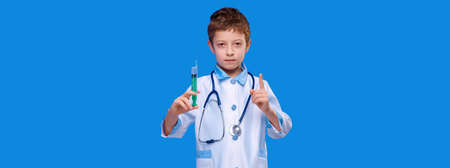 Serious boy doctor with stethoscope holds syringe and pointing up on blue isolated background. Vaccination conceptの写真素材