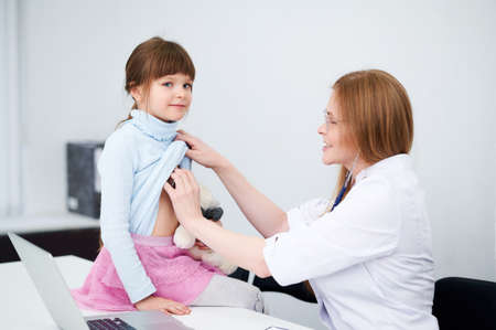 Caucasian female doctor examining patient little girl by stethoscope in officeの写真素材