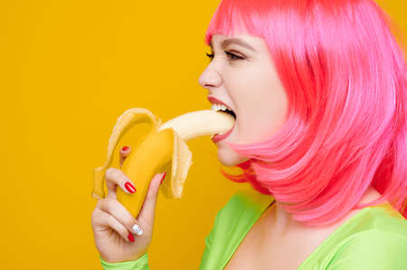 Attractive sexy hipster woman in pink wig eat banana on bright yellow backgroundの写真素材