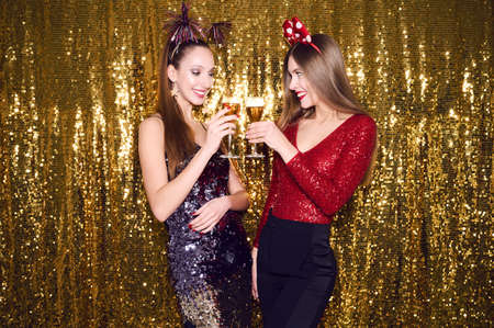Two women in fancy sparkling dresses posing isolated over vibrant gold glitter background. Positive girls hold glasses of wine spending time on happy birthday new year holiday party discothequeの写真素材