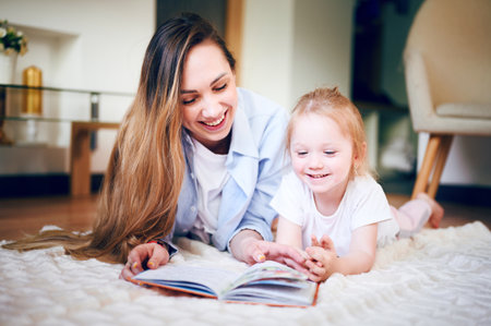 Young mother with her cute little daughter is reading book and looking at the drawings in the book while lying on the floor. The mother teaches the child. The relationship between parents and childrenの写真素材