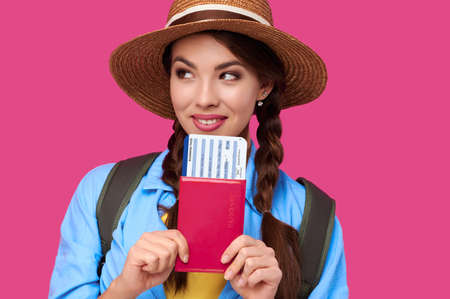 Young female traveler wearing casual clothes and straw hat holding passport with ticket on pink isolate background. Tourism concept. Studio shotの写真素材