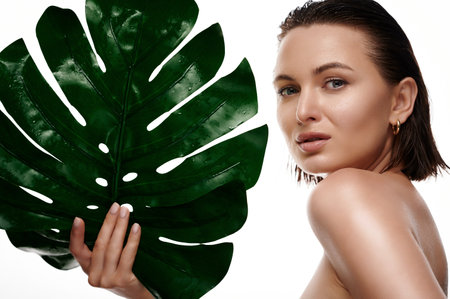 Beauty face. Middle age woman with natural makeup, healthy and wet skin holds green monstera plant isolated on white backgroundの写真素材