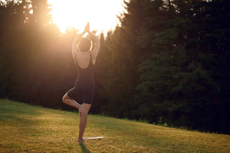 Meditate young woman standing on Sadhu nail board with closed eyes making namaste hand sign during concentration meditation practice on summer field. Concentrated female practicing yoga alone outdoorsの写真素材