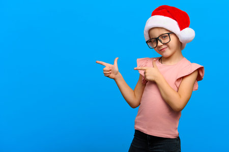 Happy new year concept. Smiling cute caucasian girl wearing glasses and santa claus red hat points at copy space on blue isolated backgroundの写真素材