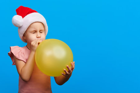 Happy new year concept. Preschool girl wearing santa claus red hat and glasses inflate yellow balloon on blue isolated backgroundの写真素材