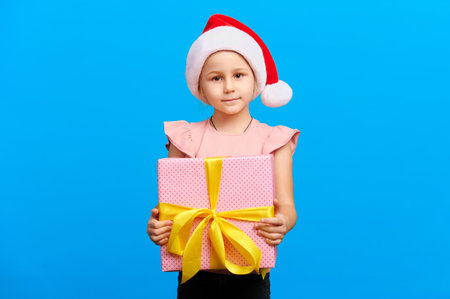 Happy new year concept. Little white girl in santa claus red hat holds christmas pink gift box with yellow bow on blue isolated background in studioの写真素材