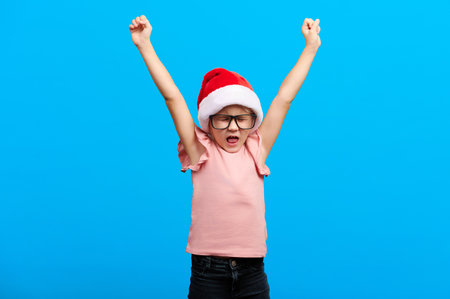 Happy new year concept. Screaming caucasian girl wearing glasses and santa claus red hat shows thumb up on blue isolated backgroundの写真素材