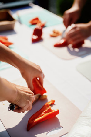 Close-up of two people cutting red bell peppers on cutting boards, with focus on their hands and the peppers. Set outdoors during a cooking activity or classの写真素材