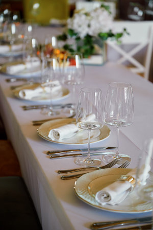 Neatly set dining table with white tablecloth, white plates, rolled napkins, wine glasses, and silverware. Elegant floral centerpiece with white flowers and green leaves in the background.の写真素材