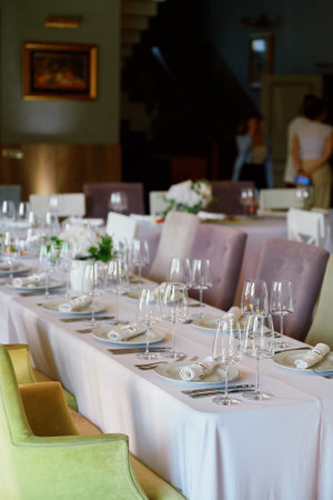 Elegant dining table set with white tablecloth, white plates, rolled napkins, wine glasses, and a...