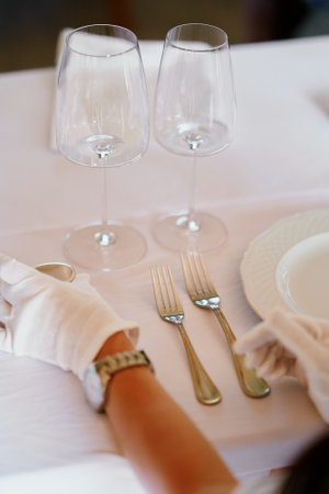 Formal dining table with a white tablecloth, white plates, silverware, two empty wine glasses, and a person wearing white gloves adjusting the settingの写真素材
