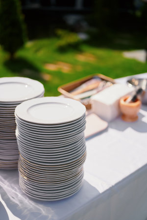 Stacks of white porcelain plates arranged neatly on a table covered with a white tablecloth. In the blurred background, a wooden tray with cutlery, napkins, and a mortar is visible. The setting is outdoors with lush green grass, suggesting a catering event or outdoor banquet.の写真素材