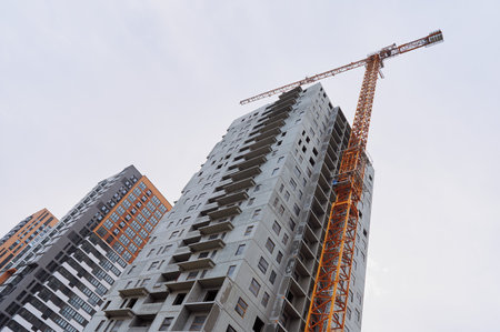 high-rise residential building under construction with exposed concrete walls and open balconies. a large yellow construction crane stands next to the unfinished skyscraper. modern apartment complex development in an urban setting with completed buildings in the backgroundの写真素材