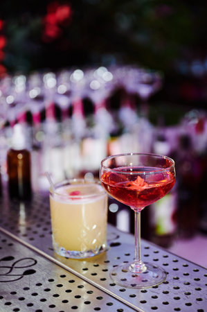 Two cocktails placed on a perforated metal bar counter. Left cocktail in a short glass with a straw and pink garnish. Right cocktail in a tall stemmed glass with pinkish-red liquid and flower garnish. Blurred bar background.の写真素材