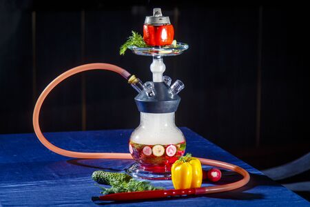 Hookah on vegetables. Hookah with a round transparent flask, which contains chopped vegetables: pepper, cucumber, radishes, herbs. Vegetables are filled with red liquor. Bowl of red pepper hookah. Lots of smoke.の写真素材
