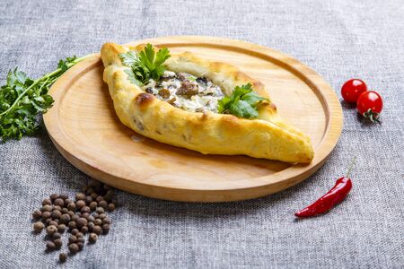 Khachapuri-boat with meat and mushrooms. Ingredients: beef tenderloin, mushrooms, cream sauce, herbs. Khachapuri is served on a round light wooden Board. photo-gray canvas. Around lies parsley, pepper, tomatoの写真素材
