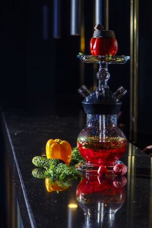 Hookah on vegetables. Hookah with a round transparent flask, which contains chopped vegetables: pepper, cucumber, radishes, herbs. Vegetables are filled with red liquor. Bowl of red pepper hookah. Lots of smoke.の写真素材