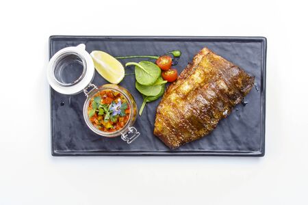 a juicy large piece of grilled fish sits on a square black plate. Next to a jar of special sauce, a...