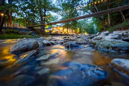 a small pedestrian suspension bridge over a shallow mountain river. Evening. Reflection of lights in the water.の写真素材