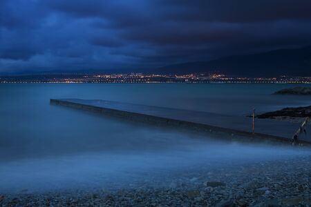 Sea surf on a long exposure in the evening. Waves roll over the pebbly beach and rocks. In the background, mountains and city lights. Long exposure. you can see a low pier that goes out to seaの写真素材