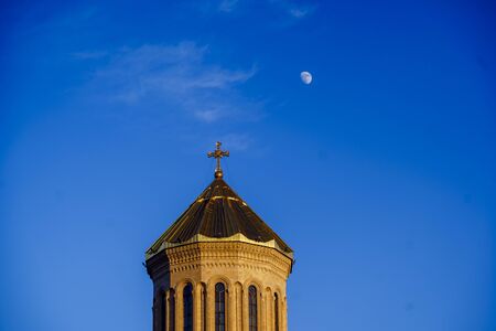 Tbilisi, winter, sunset. The dome of the Holy Trinity Cathedral (tsminda Sameba) next to the moon against the blue sky. Orange bright sunset lights up the dome and creates a festive moodの写真素材