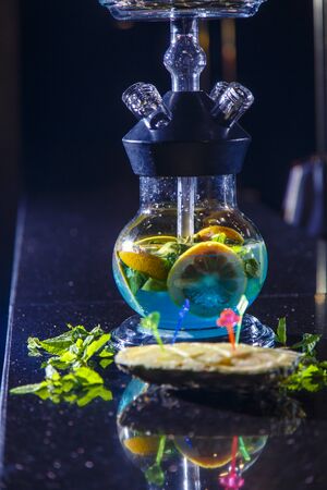 The hookah flask is round and transparent, filled with blue-green liquor. Sliced oranges and mint...