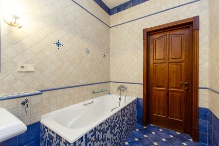 Classic rich beige bathroom with bathtub, toilet and sink with mirror. The walls are white tile with...